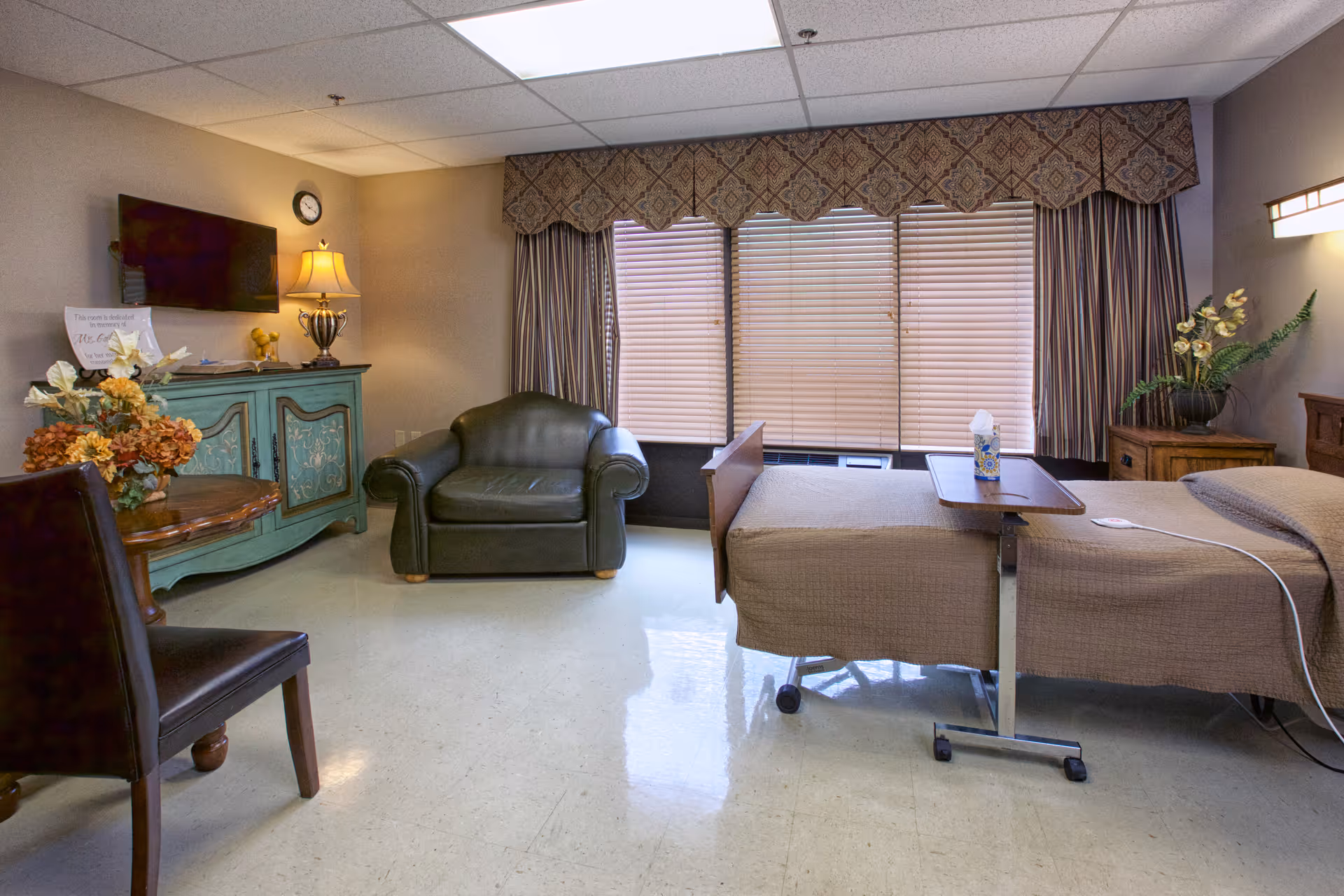 A furnished senior living bedroom with a hospital-style bed and overbed table, leather armchair, TV on a painted cabinet, and large window with blinds.