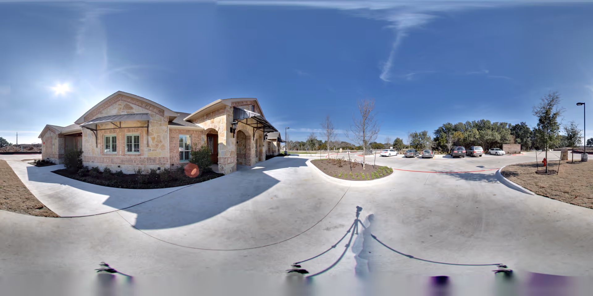 Exterior view of a single-story stone building with a covered entrance, surrounded by a concrete driveway and parking area with several parked cars. The sky is clear and blue, and there are some trees and landscaped areas around the building.