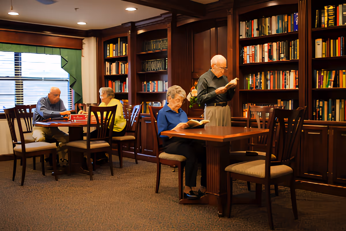 Older adults reading and playing games in a wood-paneled library/common room with bookshelves and tables.