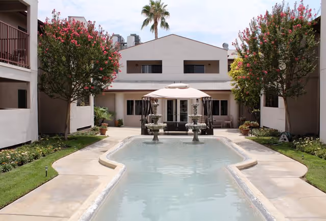 Outdoor courtyard area at Parkside Senior Apartments featuring a long rectangular water fountain with two tiered water features, surrounded by a concrete walkway. There are flowering trees with pink blossoms on either side and a building with balconies and windows in the background. A large umbrella shades seating near the fountain.