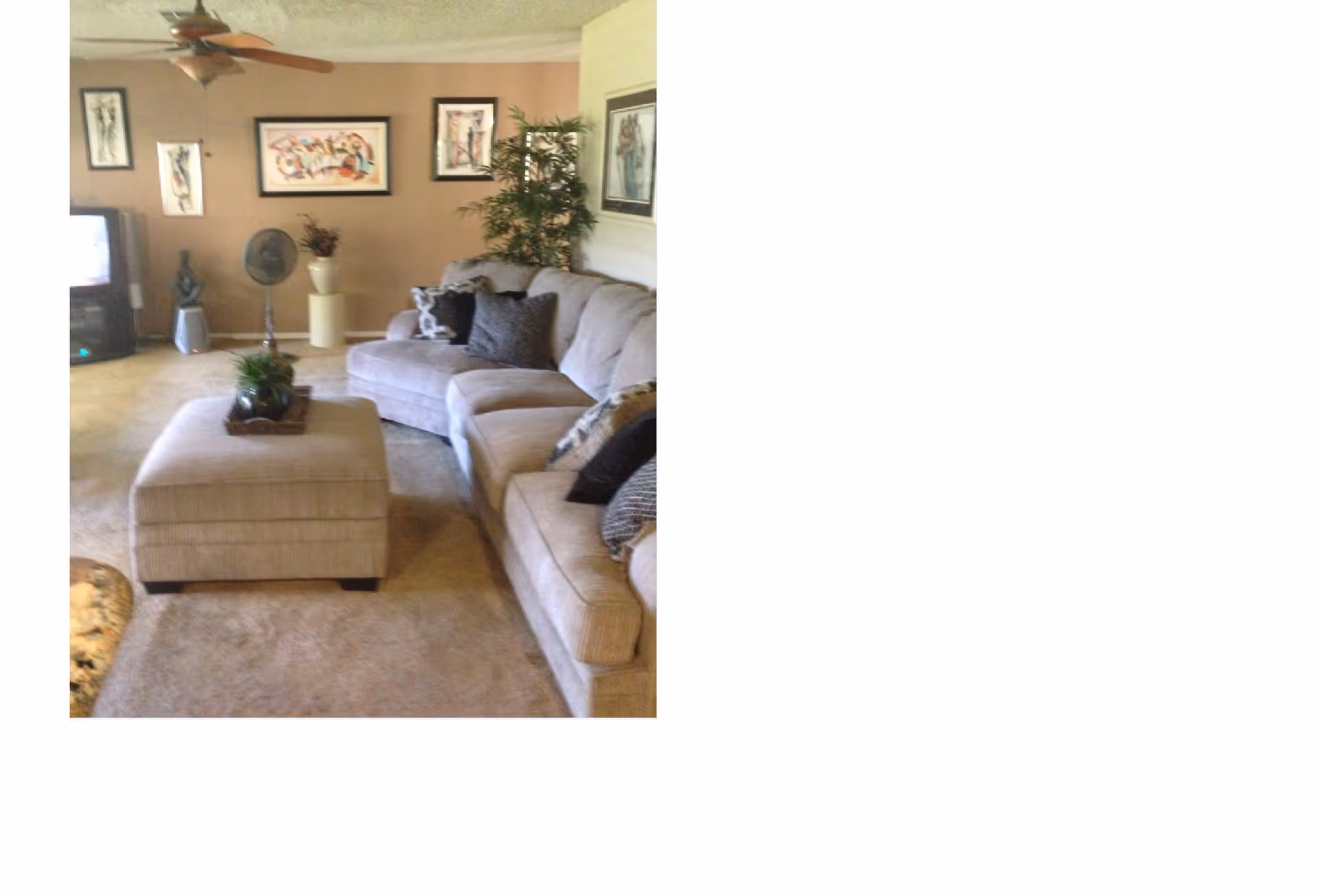 A cozy living room with a large beige sectional sofa adorned with multiple throw pillows. In front of the sofa is a matching ottoman with a small plant on top. The room has beige carpet and walls decorated with framed artwork. A ceiling fan is visible, and there is a TV in the corner along with a standing fan and some decorative items.