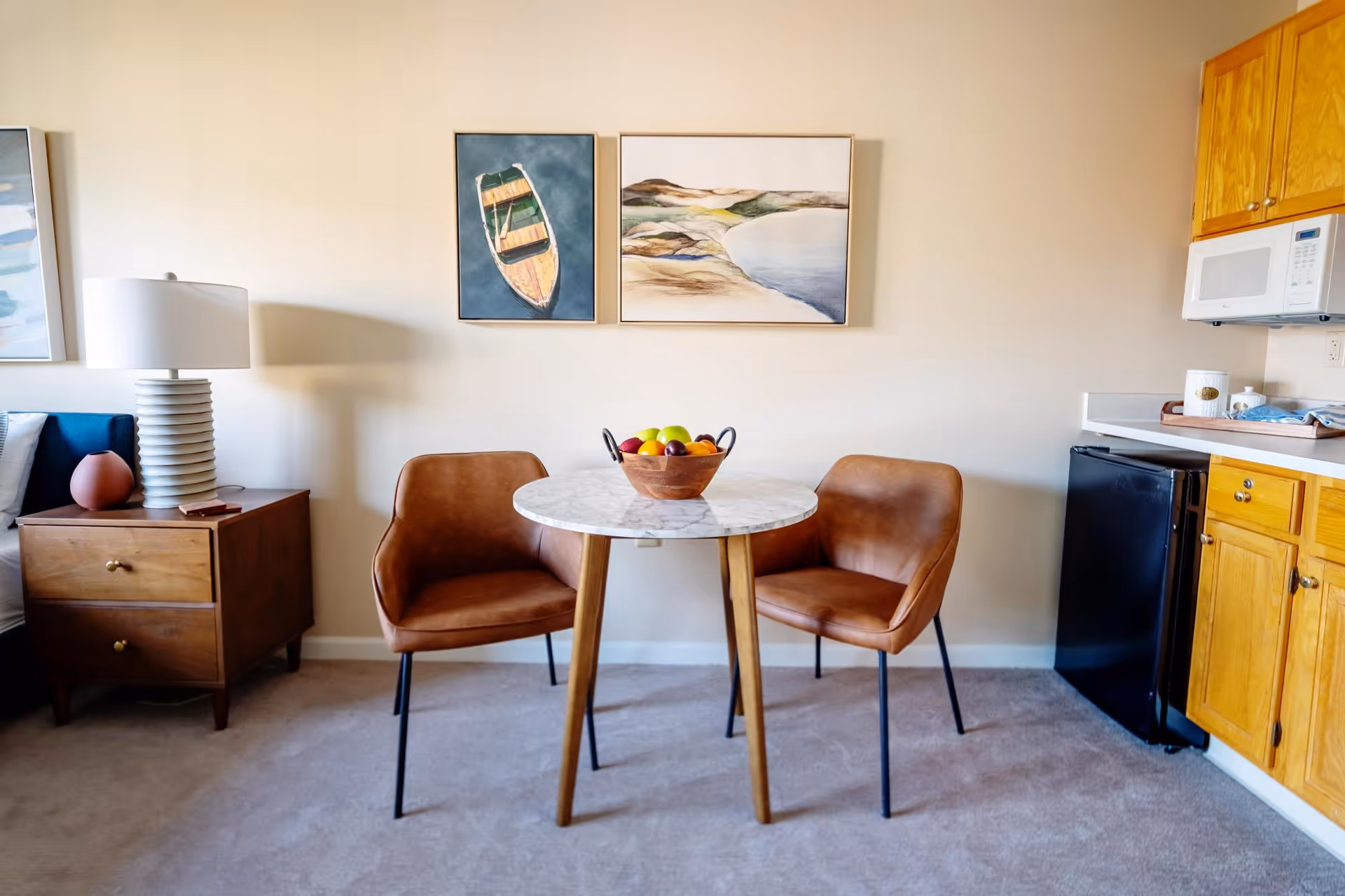 Small dining area with a round marble table, two brown chairs, a fruit bowl, and kitchenette cabinets to the right.