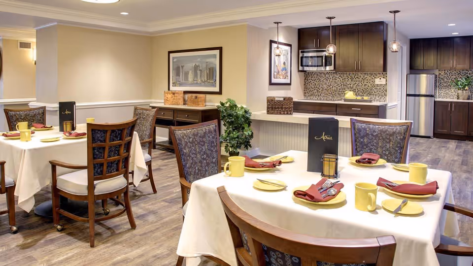 A dining area in a senior living facility with two tables covered in white tablecloths, each set with yellow plates, yellow mugs, and red napkins. The chairs have wooden frames and patterned upholstery. In the background, there is a kitchen area with dark wood cabinets, a microwave, a refrigerator, and a mosaic tile backsplash. The walls are painted beige, and there is a framed picture and a plant near the kitchen counter.
