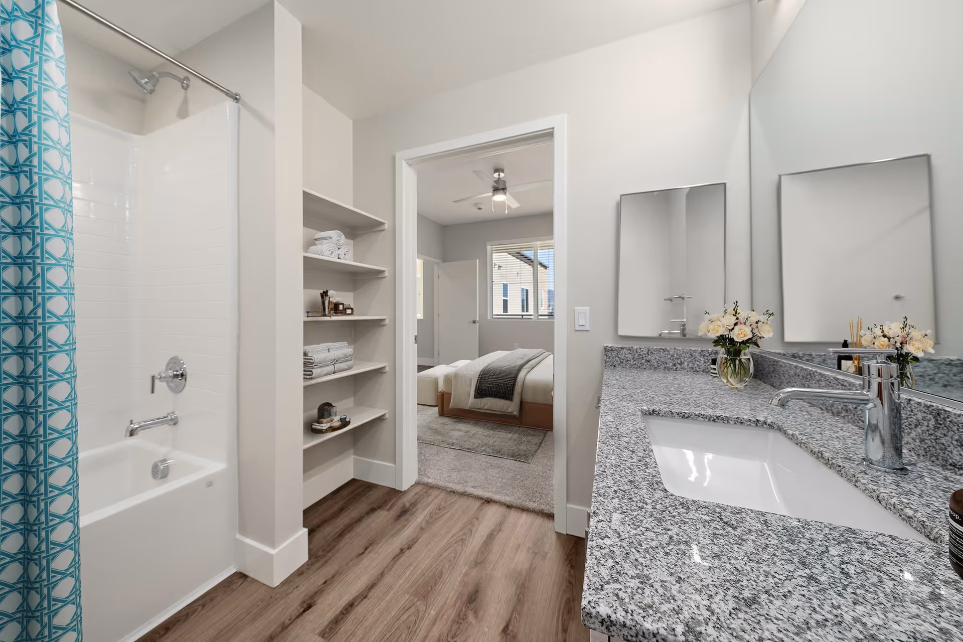 A modern bathroom with a granite countertop and sink on the right, a large mirror above the sink, and a vase with flowers on the counter. On the left, there is a bathtub with a blue patterned shower curtain and a built-in shelving unit holding towels and toiletries. Through an open doorway, a bedroom with a bed, ceiling fan, and window is visible.