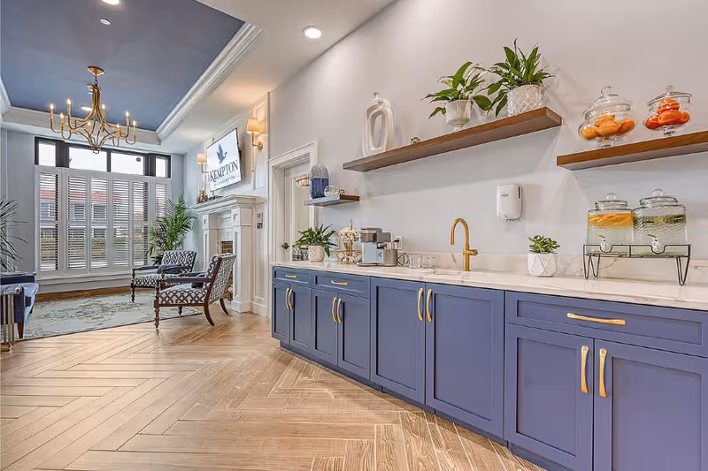 A bright and elegant interior space featuring a long counter with blue cabinets and gold handles, a gold faucet, and two beverage dispensers with infused water. Above the counter are two wooden floating shelves decorated with plants and glass jars. In the background, there is a seating area with two patterned armchairs, a fireplace with a mounted TV displaying 'The Kempton of Charleston,' and large windows with white shutters letting in natural light. The ceiling has a blue accent with a gold chandelier, and the floor is a light wood herringbone pattern.