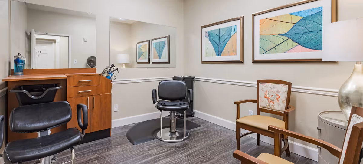 Interior view of a small salon or barber area in a senior living facility. The room features a black salon chair on a protective mat, a wooden cabinet with drawers, a large wall mirror, two framed colorful leaf prints on the wall, two wooden chairs with cushioned seats and backs, and a lamp on a small round table.