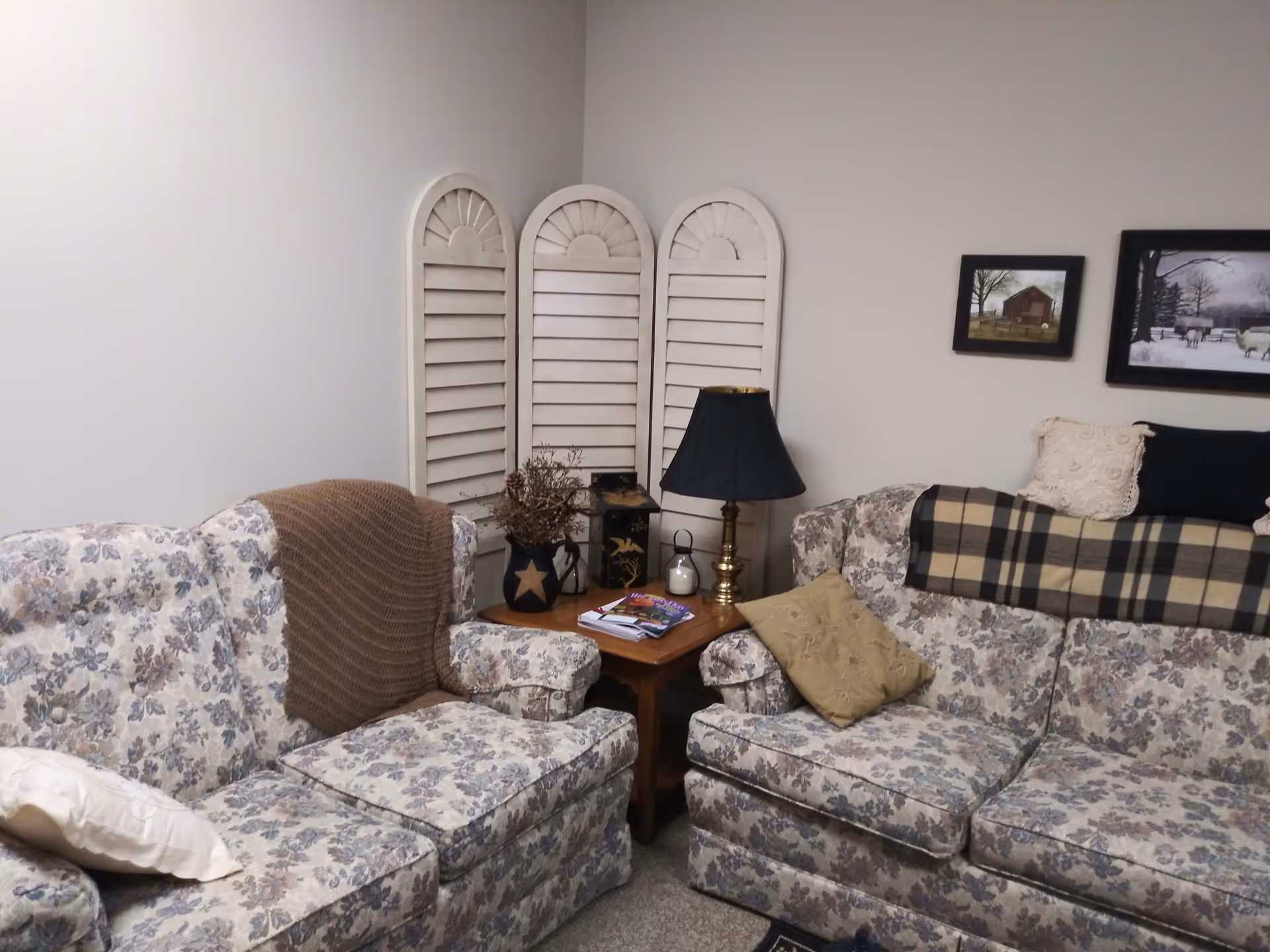 A cozy living room corner with two floral-patterned sofas adorned with various pillows and throws. Between the sofas is a wooden side table holding a black lampshade, decorative items, and magazines. Behind the table, there is a white three-panel shutter-style room divider. Two framed pictures hang on the wall above one of the sofas.