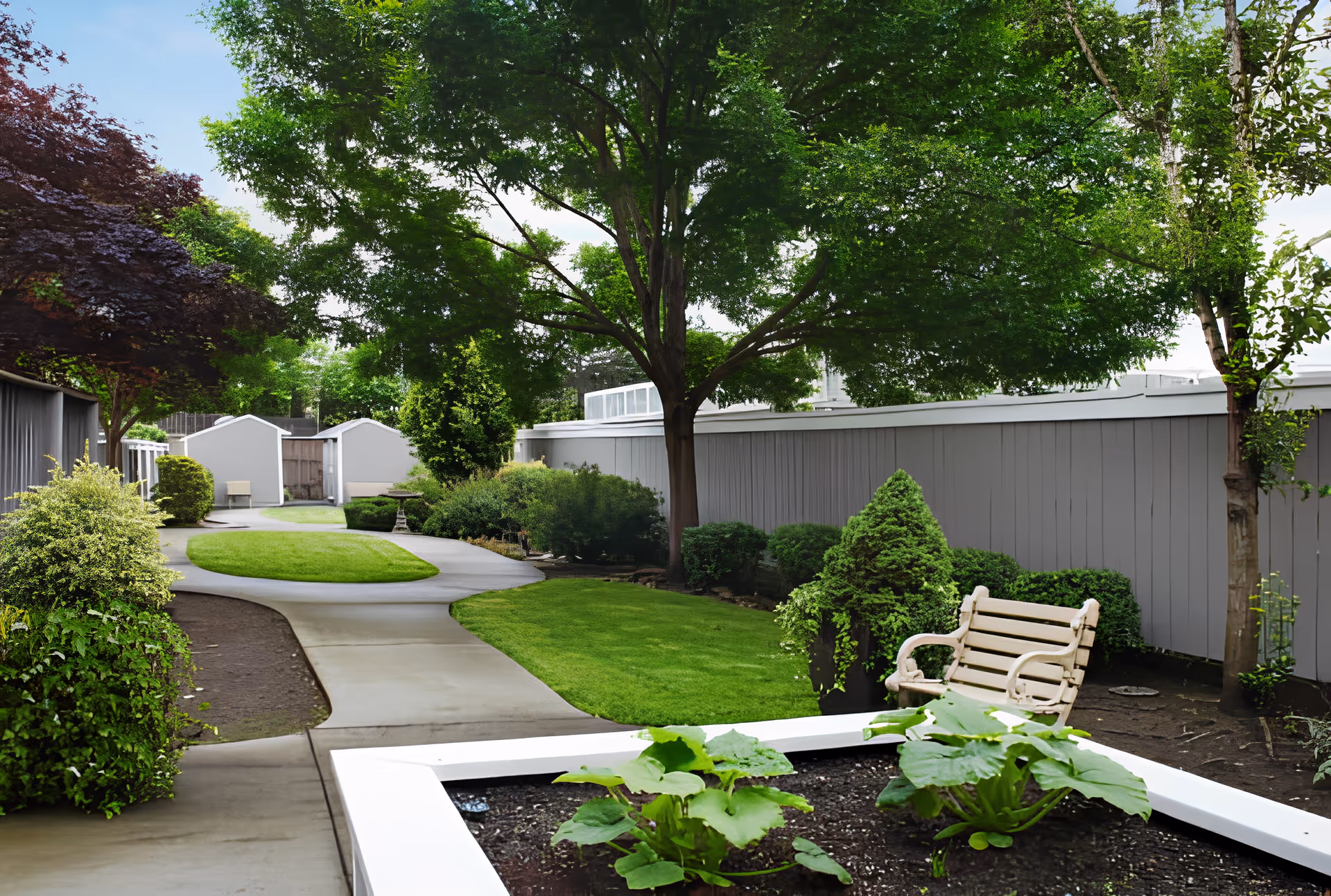 A peaceful outdoor garden area with a winding concrete pathway, green grass, various bushes and trees, a white wooden bench, and a raised garden bed with plants. The area is enclosed by a tall gray fence and has a clear blue sky above.