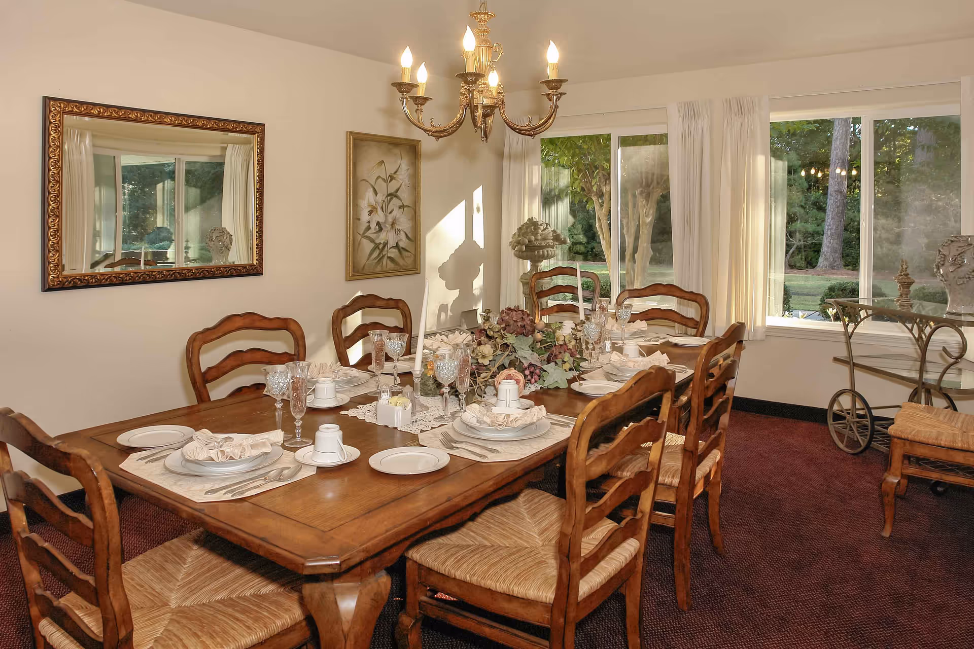 A dining room with a wooden table set for six people, featuring plates, glasses, napkins, and candles. The room has wooden chairs with woven seats, a chandelier hanging above the table, a large mirror on one wall, a floral painting, and large windows with white curtains showing a green outdoor view.