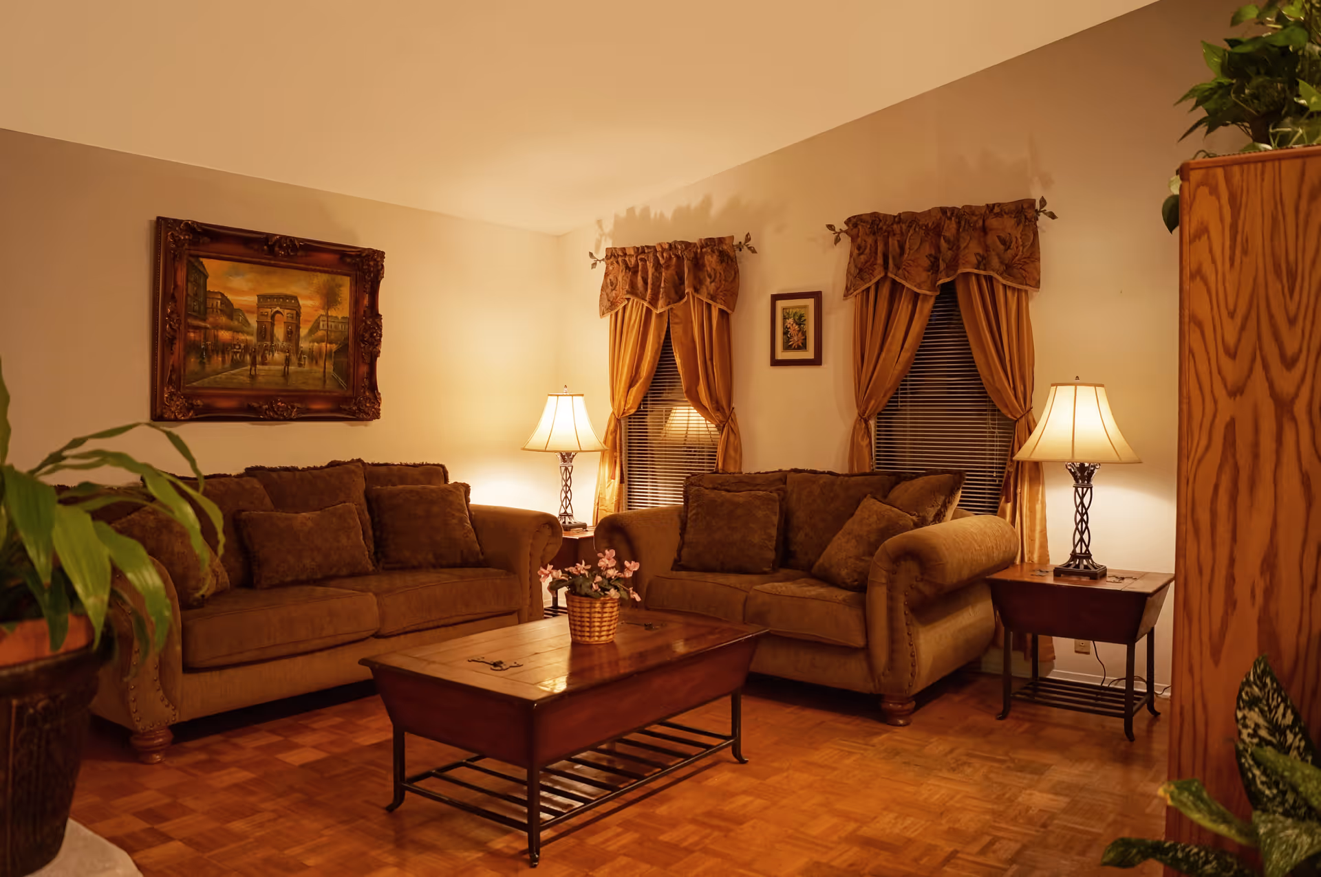 A cozy living room with two brown sofas, a wooden coffee table with a small plant, two side tables each with a lamp, framed artwork on the walls, and windows with brown curtains. There are also some green plants visible in the corners of the room.