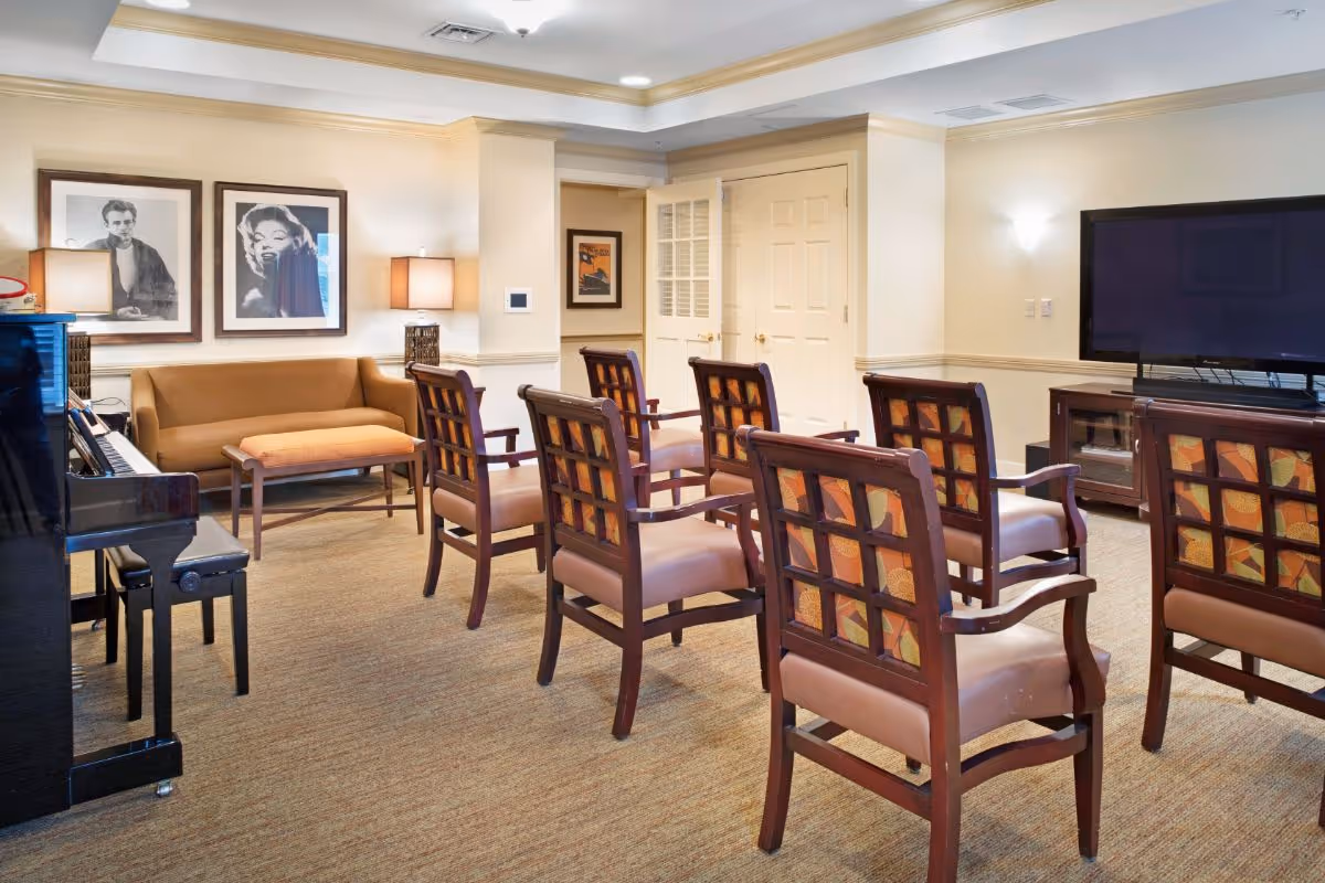 A well-lit common room with several wooden chairs arranged in rows facing a large flat-screen TV on a wooden stand. To the left, there is a piano with a bench and a brown sofa with an ottoman in front of it. The walls are decorated with framed black and white photographs and a lamp on a side table.