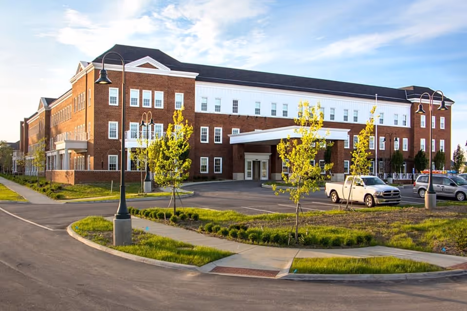 Front exterior of a three-story brick senior living facility with a covered entrance, parking lot, and young trees.