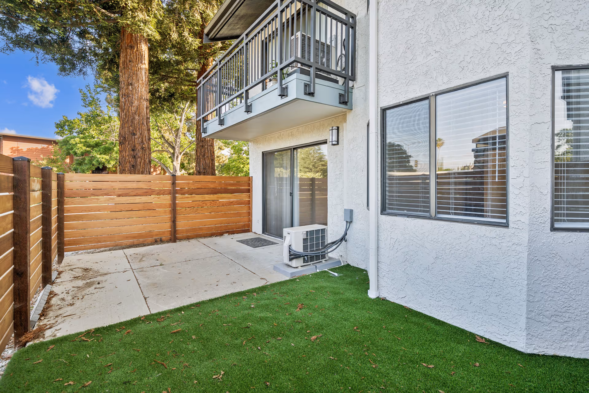 Outdoor patio area with artificial grass, concrete flooring, a wooden privacy fence, and a balcony above. The exterior wall of the building is white with two windows and a sliding glass door. There are tall trees visible in the background.