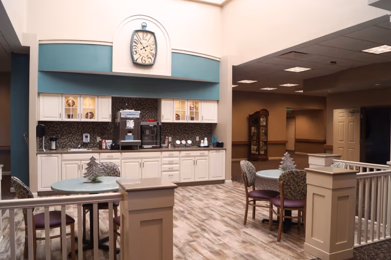 Interior view of a senior living facility common area with a kitchenette featuring white cabinets, a coffee machine, and a large decorative clock above. There are two round tables with chairs around them, each table decorated with a small silver Christmas tree. The flooring is wood-patterned, and the area is well-lit with ceiling lights. A grandfather clock is visible in the background near a hallway.