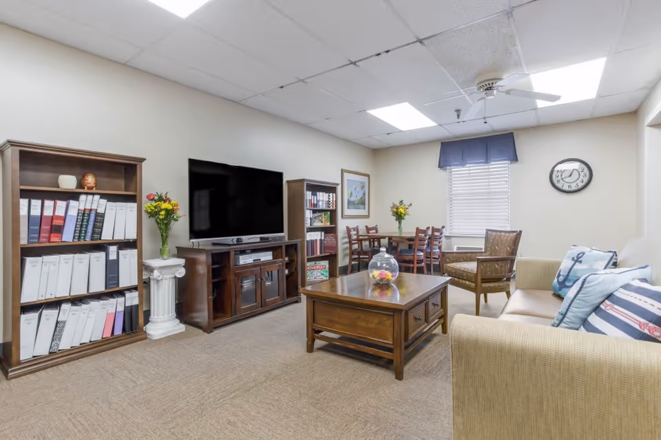 A cozy living room area in a senior living facility featuring a beige sofa with nautical-themed pillows, a wooden coffee table with a glass bowl centerpiece, a wooden TV stand with a flat-screen television, bookshelves filled with binders and books, a small dining table with chairs near a window with blinds and a blue valance, a wall clock, and a ceiling fan with lights.