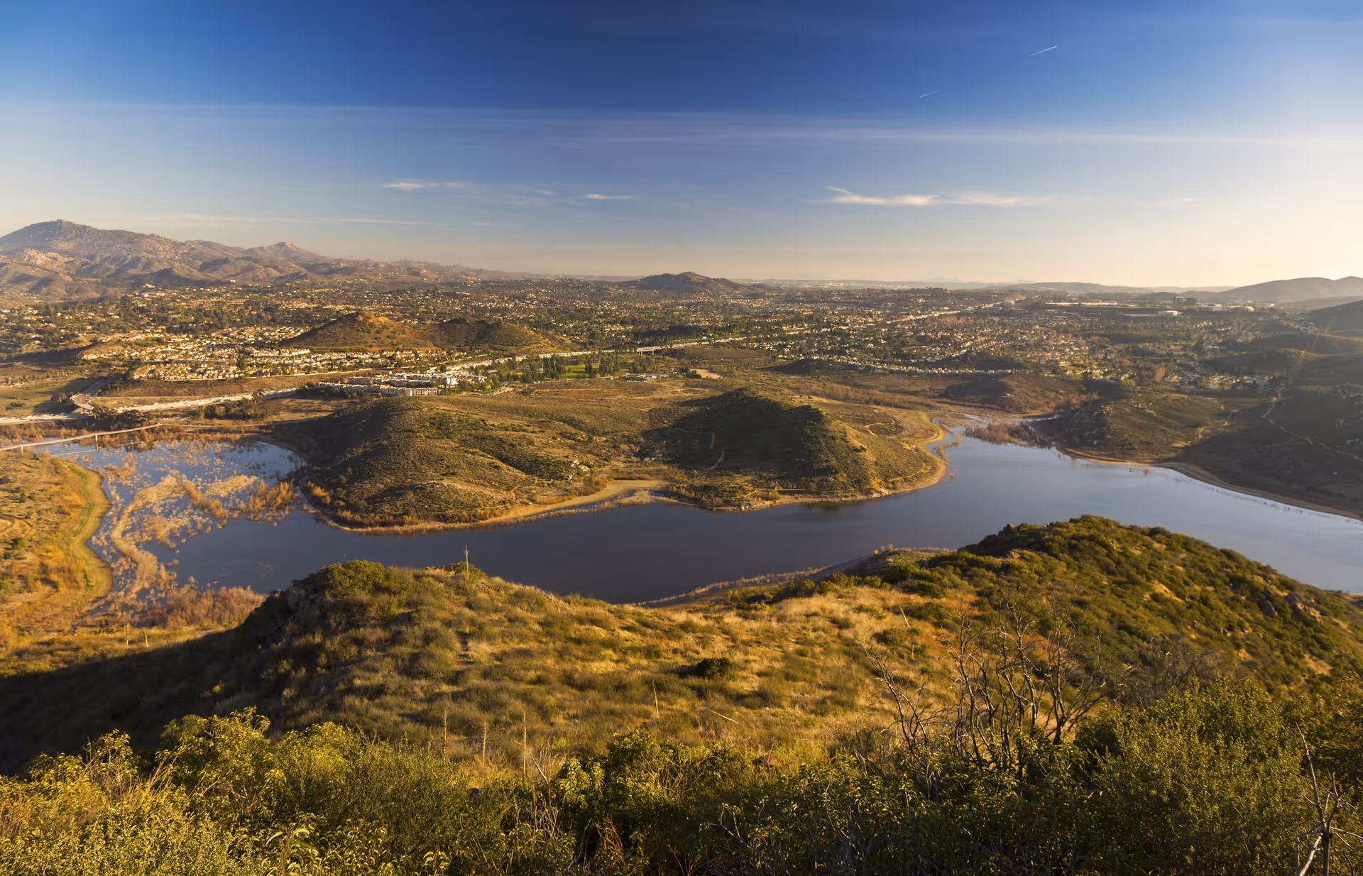 Aerial view of a winding reservoir surrounded by rolling hills and distant suburbs under a clear sky.