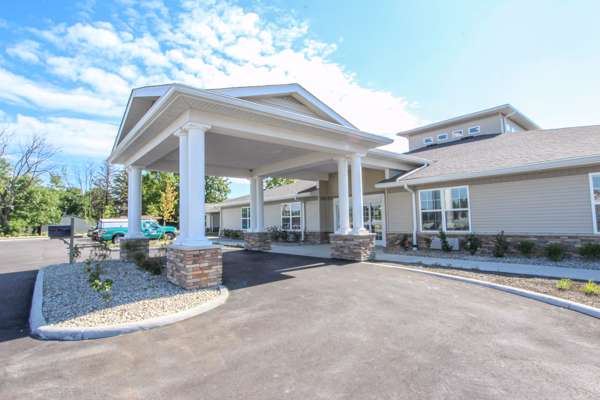 Exterior view of Lincolnshire Place - Memory Care & Assisted Living showing the entrance with a covered drop-off area supported by white columns with stone bases. The building is light-colored with multiple windows and a clear blue sky with some clouds above.