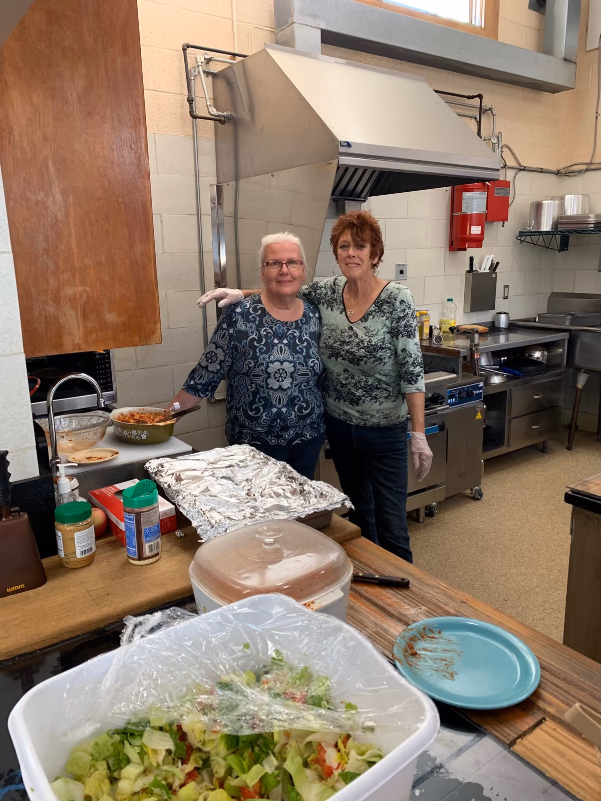 Two women wearing gloves stand in a commercial kitchen behind a counter with a large salad, covered dishes, and cooking equipment.
