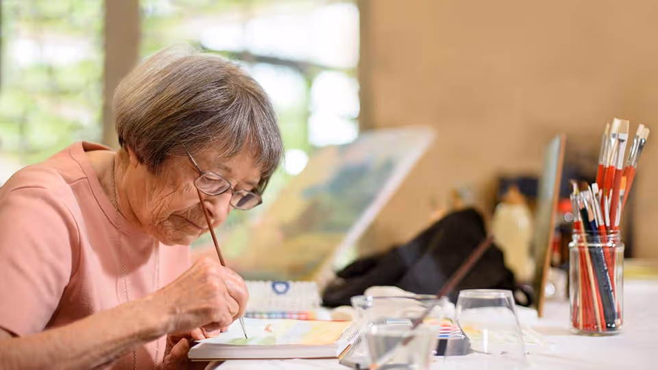 An elderly woman wearing glasses and a pink shirt is focused on painting with a brush on a canvas or paper at a table. Various painting supplies, including brushes in a jar and watercolor paints, are visible on the table. The background shows a blurred window with greenery outside.