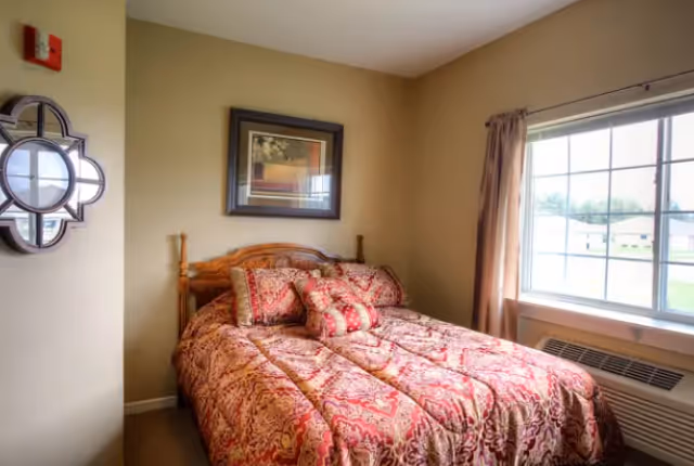 A cozy bedroom with a wooden bed frame and a red patterned comforter and pillows. There is a window with beige curtains letting in natural light, a framed picture on the wall above the bed, and a decorative round mirror on the adjacent wall.