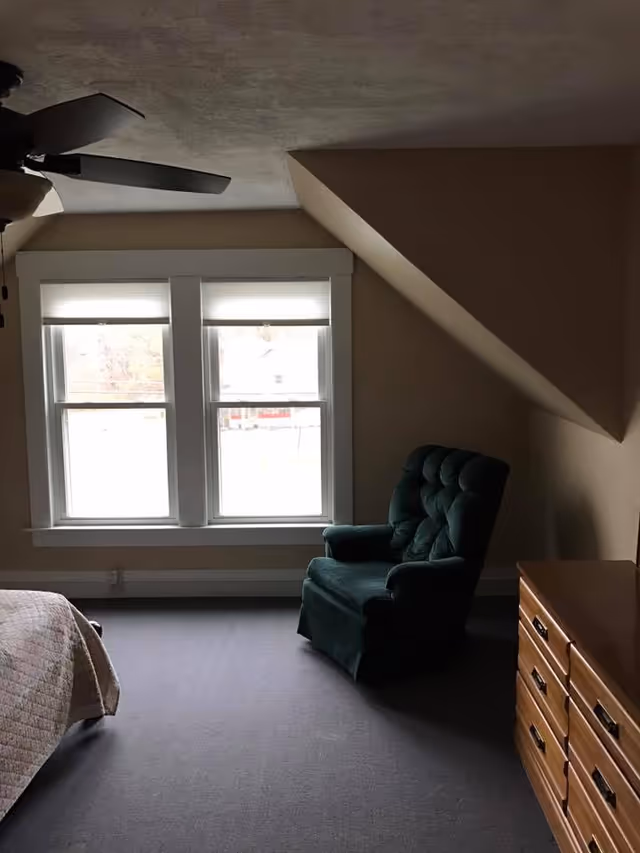 A bedroom with a slanted ceiling, featuring a green upholstered armchair near two large windows with white blinds. There is a wooden dresser with multiple drawers on the right side and part of a bed with a quilted bedspread visible on the left. A ceiling fan is partially visible at the top left corner.