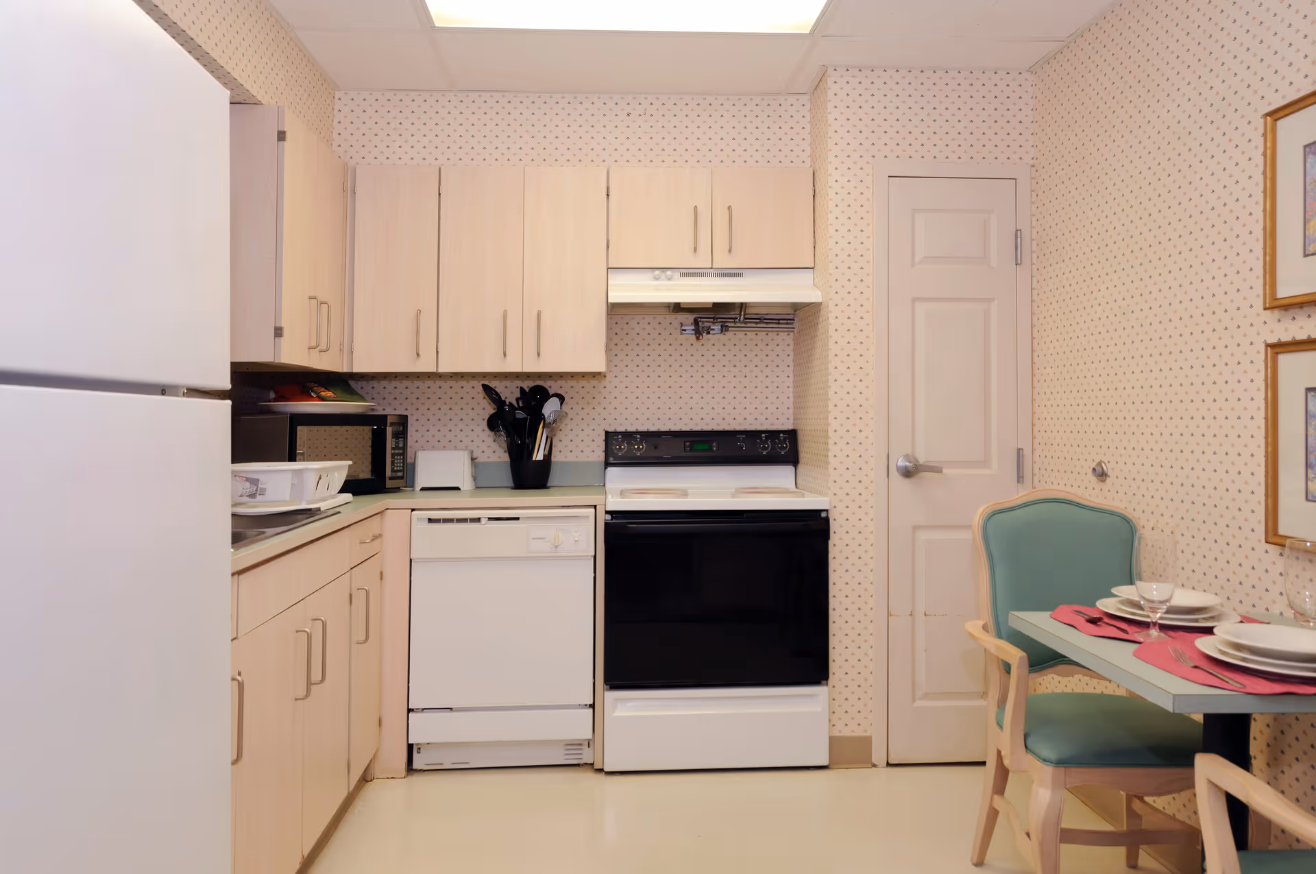 Compact kitchen with white refrigerator, dishwasher and stove, light wood cabinets, and a small dining table with a teal chair.
