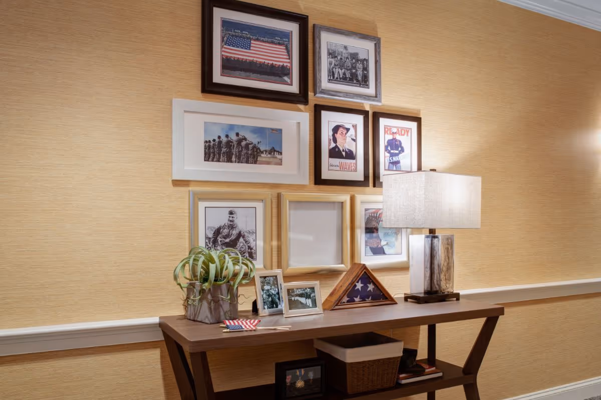 A wooden console table against a beige textured wall with a collection of framed photographs and artwork above it. On the table are a potted plant, two small framed photos, a folded American flag in a display case, a table lamp with a rectangular shade, and a basket on the lower shelf. The photos and artwork include military and patriotic themes.