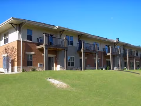 Exterior view of a two-story apartment building with balconies on the upper floor and a grassy lawn in front under a clear blue sky.