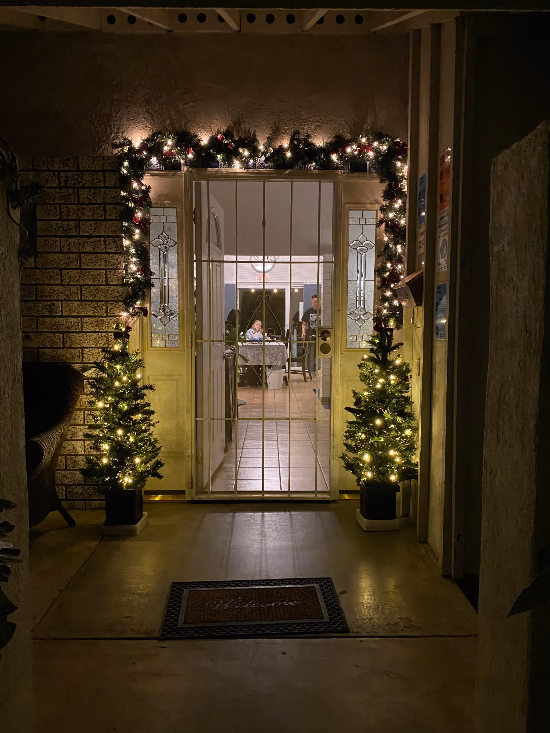 View through a decorated entrance door with Christmas garlands and two small lit Christmas trees on either side. Inside, a dining area is visible with people sitting at a table.
