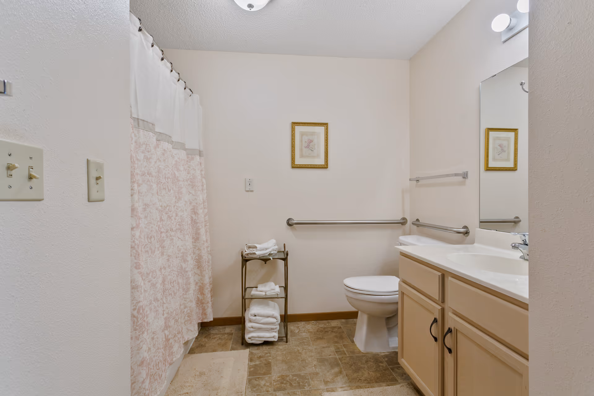 A clean bathroom with a shower curtain on the left, a toilet with grab bars on the wall behind it, a small metal rack holding folded towels, and a vanity with a sink and mirror on the right. The walls are light-colored and there is a framed picture hanging above the toilet.