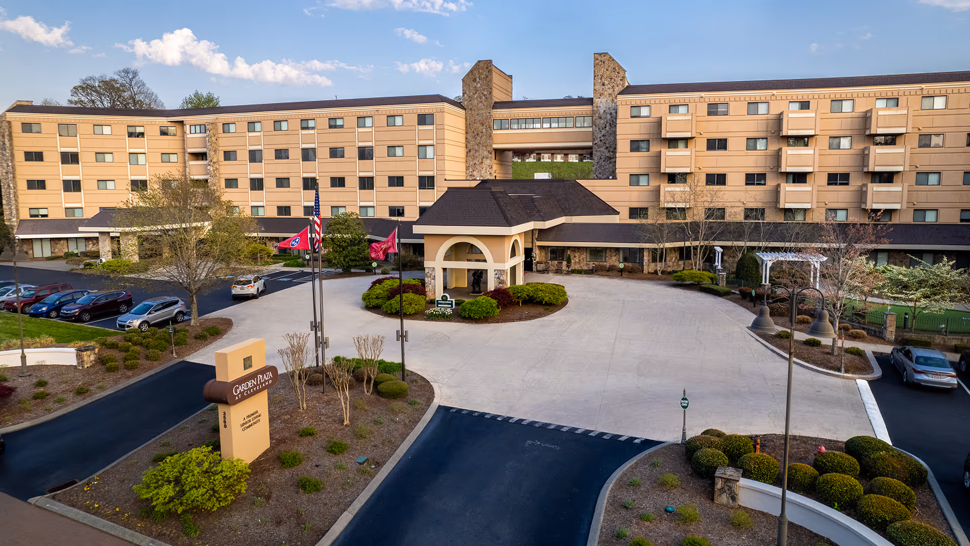 Front entrance of a multi-story senior living building with a covered porte-cochère, circular driveway, flagpoles, landscaping and a sign reading Garden Plaza.