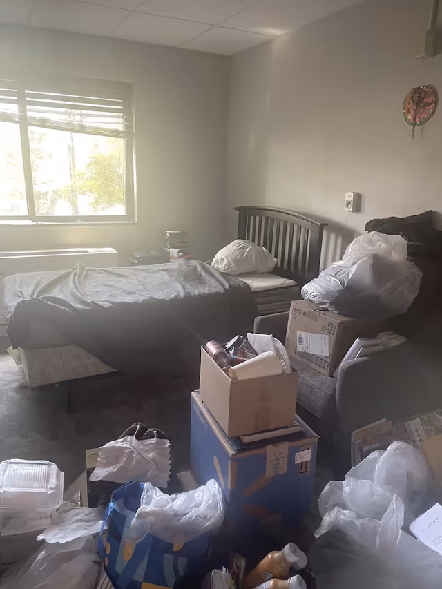 A cluttered bedroom with a single bed against the wall under a window. The bed has a gray blanket and a white pillow. There is a gray armchair next to the bed piled with boxes and plastic bags. The floor is covered with various bags, boxes, and miscellaneous items. The walls are plain and light-colored, with a small decorative item hanging on the right wall.