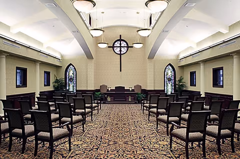 Interior chapel with rows of chairs facing a pulpit, stained-glass windows, and a prominent wall cross.