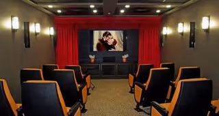 Small theater room with rows of black and orange cushioned seats facing a large screen displaying a couple embracing. The room has beige walls with wall-mounted lights and red curtains framing the screen.