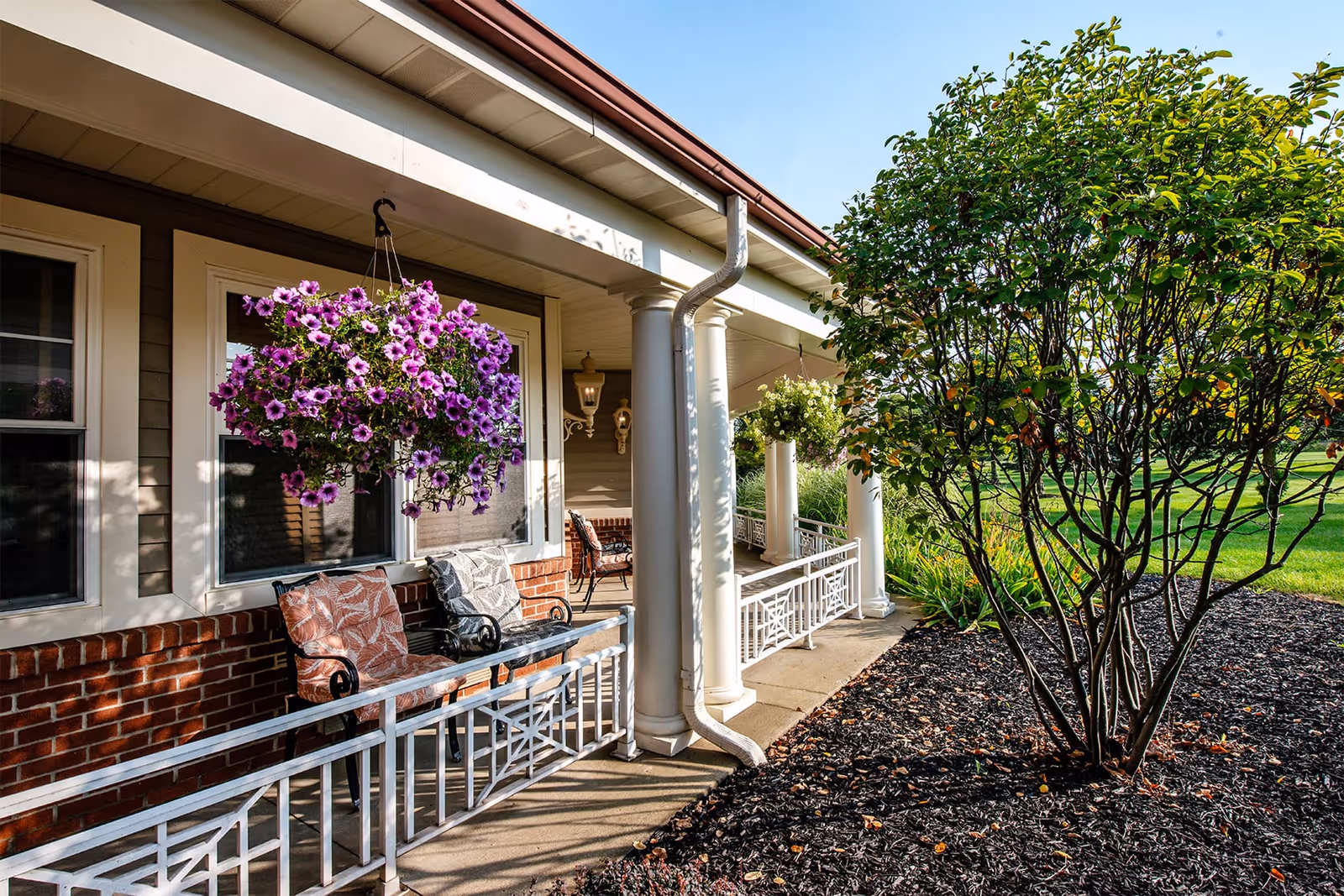 A porch area of a building with white columns and a white railing. There are two cushioned chairs with patterned pillows, hanging baskets with purple and white flowers, and a tree with green leaves in a mulched garden bed. The scene is well-lit with sunlight and shows a green lawn in the background.