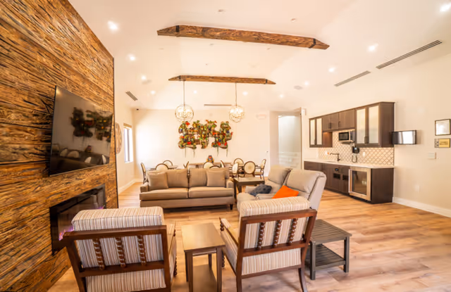 A spacious and well-lit living room area featuring a large flat-screen TV mounted on a rustic wooden accent wall with a fireplace below. The room includes a beige sofa, two striped armchairs, a coffee table, and a side table. In the background, there is a dining area with a round table and chairs, decorative wall art, and a kitchenette with dark cabinets, a microwave, and a small refrigerator. The ceiling has exposed wooden beams and modern pendant lights.