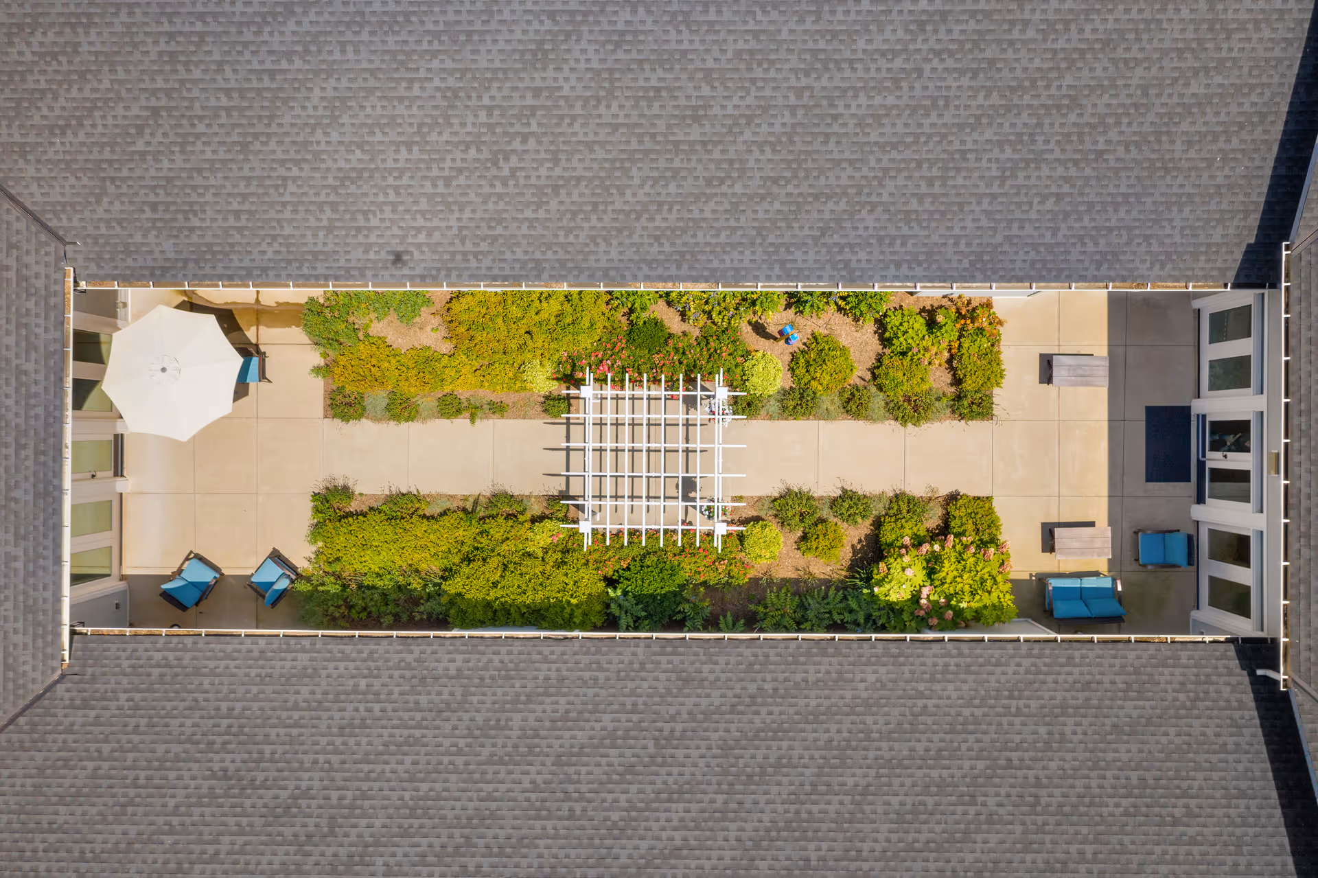 Aerial view of a rectangular courtyard garden enclosed by a building roof. The courtyard features a central pathway with a white pergola, surrounded by green shrubs and plants. There are seating areas with blue chairs and tables, and a white patio umbrella on one side.