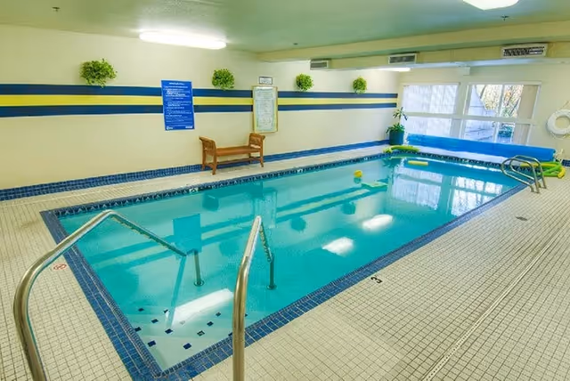 Indoor swimming pool with clear blue water, metal handrails for entry, tiled floor and walls, a wooden bench against the wall, potted plants, and large windows letting in natural light.