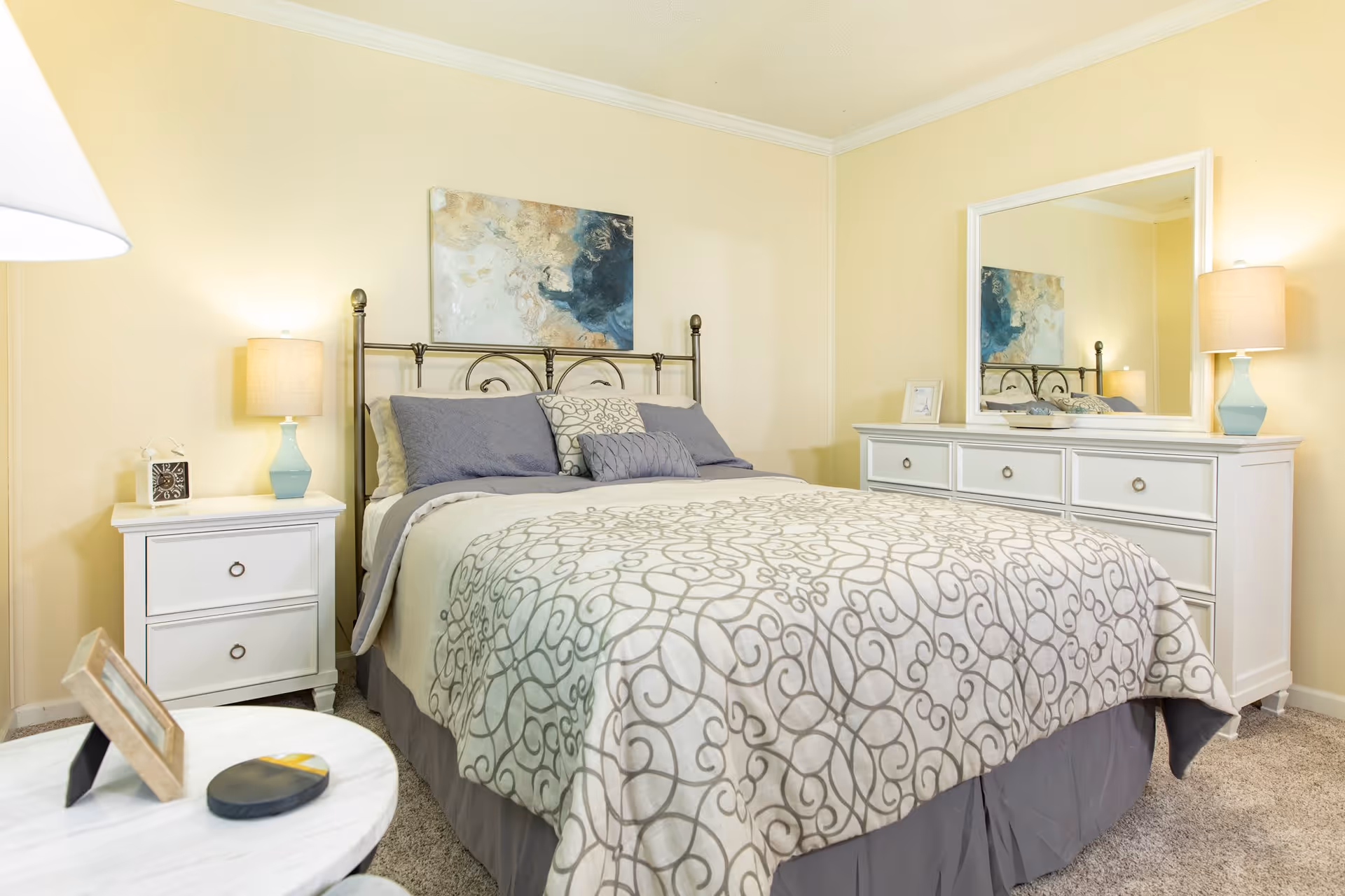 A cozy bedroom with a neatly made bed featuring a patterned comforter and multiple pillows. On either side of the bed are white nightstands with lamps. A large white dresser with a mirror above it is positioned against the wall. The walls are painted a soft yellow, and there is a decorative painting above the bed.