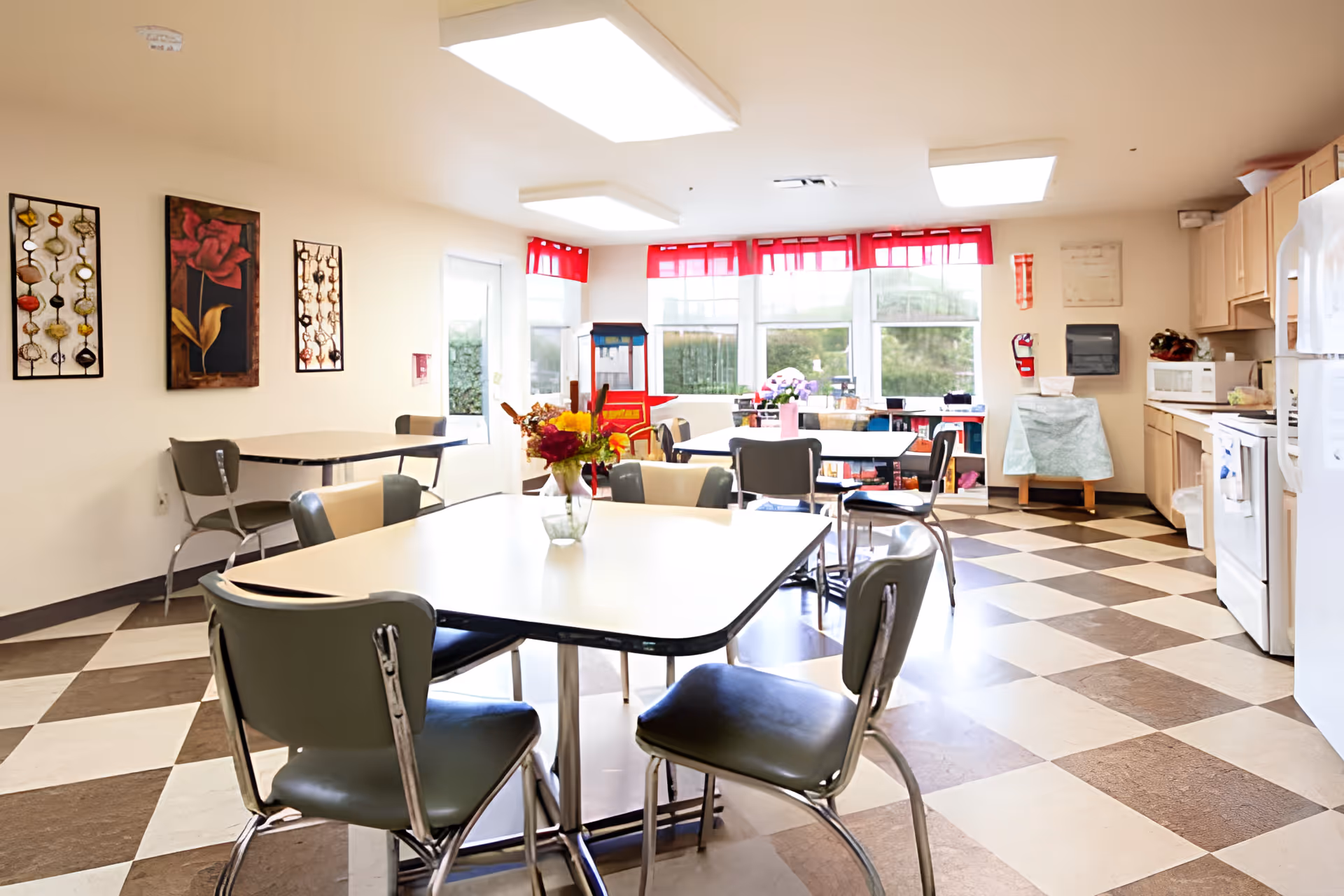 A bright and clean dining area in a senior living facility with checkered brown and white flooring, several tables with chairs around them, and a small vase of flowers on the nearest table. The room has large windows with red valances letting in natural light, wall art, and a kitchenette area with appliances including a refrigerator, microwave, and stove.