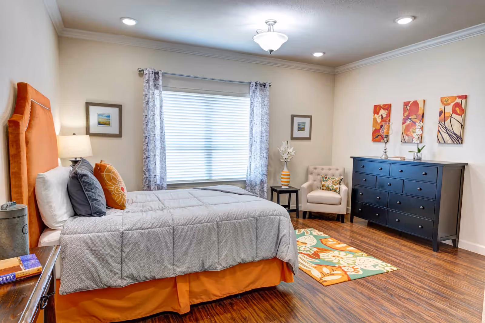 A bright and cozy bedroom with a large bed featuring an orange headboard and gray bedding. The room has wooden flooring, a window with patterned curtains, a beige armchair with a floral cushion, a black dresser with multiple drawers, and colorful floral artwork on the wall above the dresser. A small side table with a striped vase and white flowers is next to the armchair.