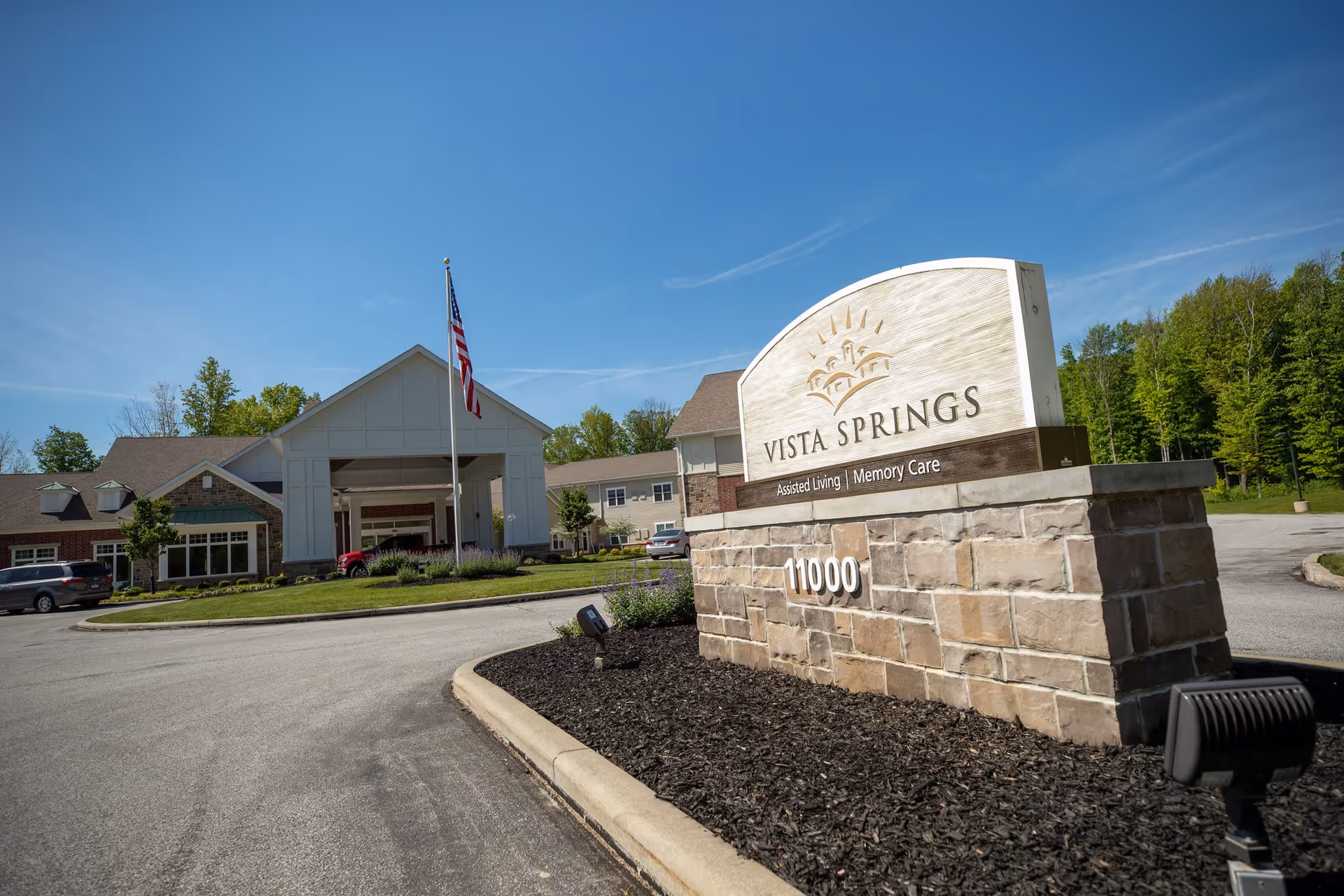Entrance to Vista Springs senior living facility with a large stone sign displaying the name and address, an American flag on a pole, and a building with a covered driveway surrounded by greenery under a clear blue sky.