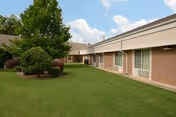 Exterior view of a single-story brick building with multiple windows and a well-maintained lawn with trimmed bushes and a tree under a partly cloudy sky.