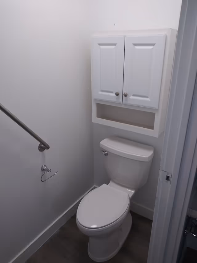 A small bathroom with a white toilet, a white wall-mounted cabinet with two doors above the toilet, a metal handrail on the left wall, and a toilet paper holder without a roll.