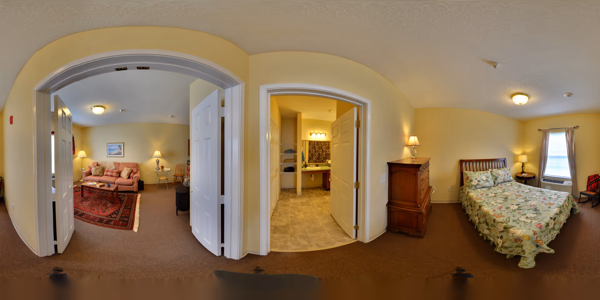 Interior view of a senior living facility apartment showing a living room with a sofa, coffee table, and lamps on the left, a bathroom with a sink and mirror in the center, and a bedroom with a bed, nightstand, and window on the right. The walls are painted light yellow and the floors are carpeted except for the bathroom which has tile flooring.