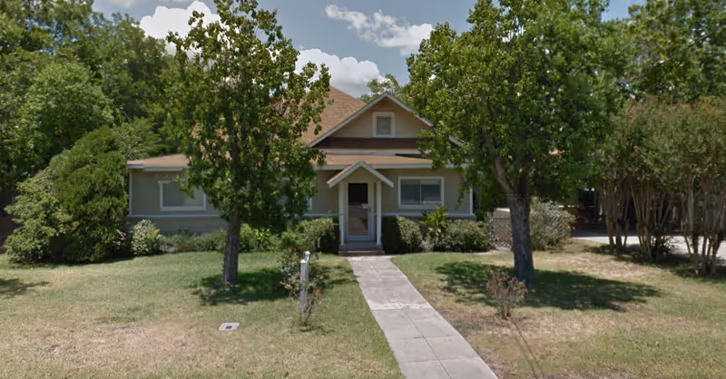 Front exterior view of a single-story house with a gable roof, surrounded by trees and bushes, with a concrete walkway leading to the front door.