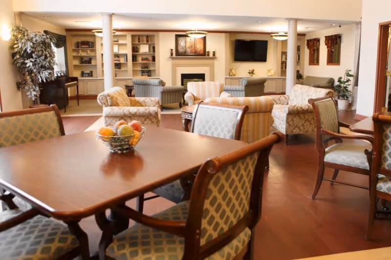 Communal sitting and dining area with a wooden table and upholstered chairs in the foreground and sofas arranged around a fireplace and TV in the background.
