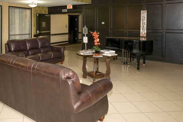 Lobby-style seating area with leather sofas, a round table with flowers and magazines, and a grand piano against a dark paneled wall.