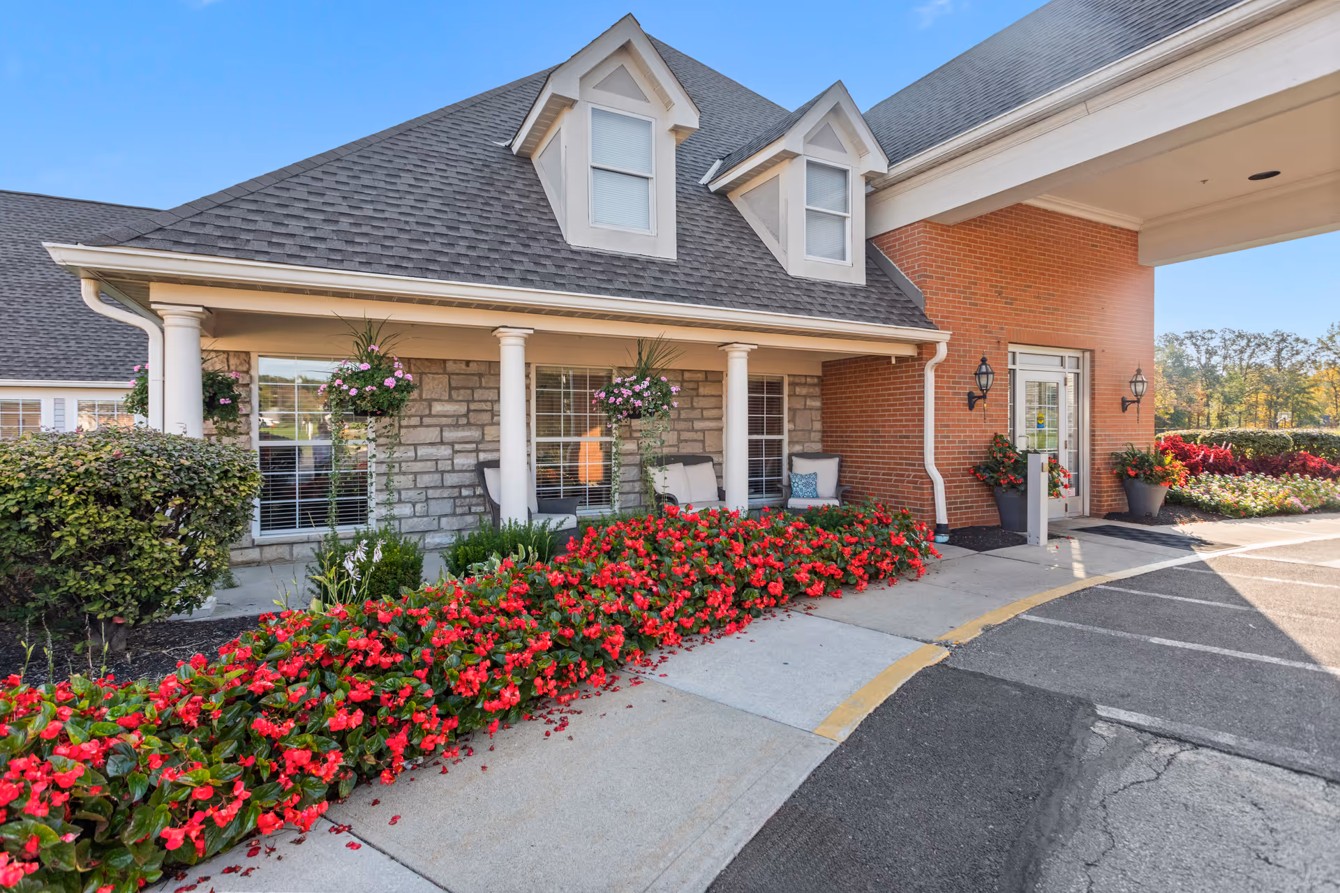 Exterior view of The Gables of Marysville senior living facility entrance with a covered porch, white columns, hanging flower baskets, red flowers along the sidewalk, and a brick and stone facade under a clear blue sky.