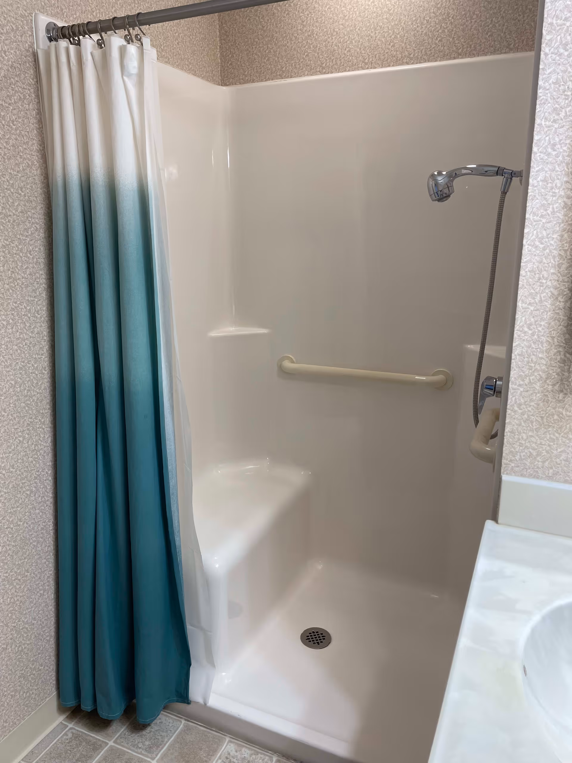 A clean shower area with a white built-in seat and grab bar. The shower has a handheld showerhead attached to the wall and a blue and white gradient shower curtain. The surrounding walls have a light patterned wallpaper, and part of a white sink countertop is visible on the right.